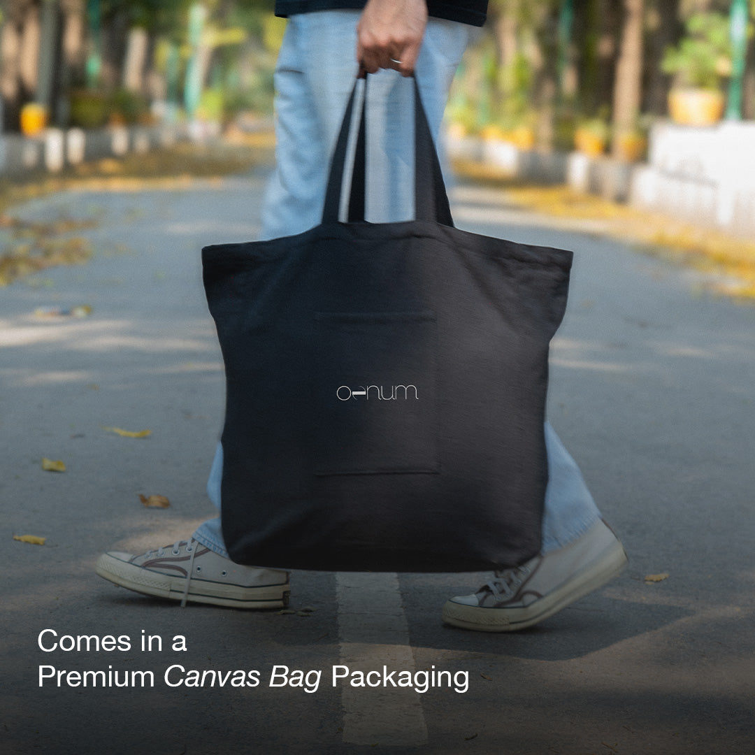 Product packaging photo for OENUM Plasma car cover, showing a person's legs and hand carrying a black canvas tote bag with long handles and the 'oenum' logo printed in white on the front pocket, walking along a tree-lined outdoor path with fallen leaves; overlay text reads 'Comes in a Premium Canvas Bag Packaging.